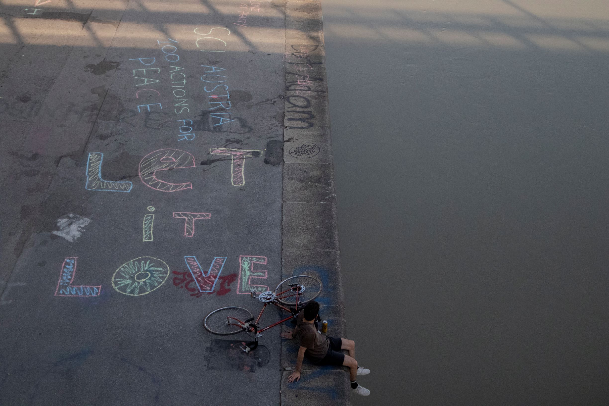 a man sitting by the river with a bike, on one half can be seen the river, in the other is the sidewalk with drawings