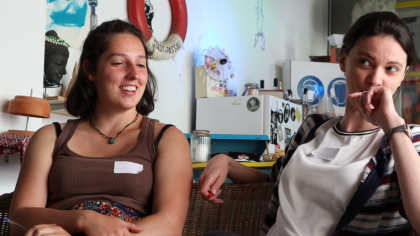 two woman smiling, in the bckground there is a fridge, a wall with decorations, a jar